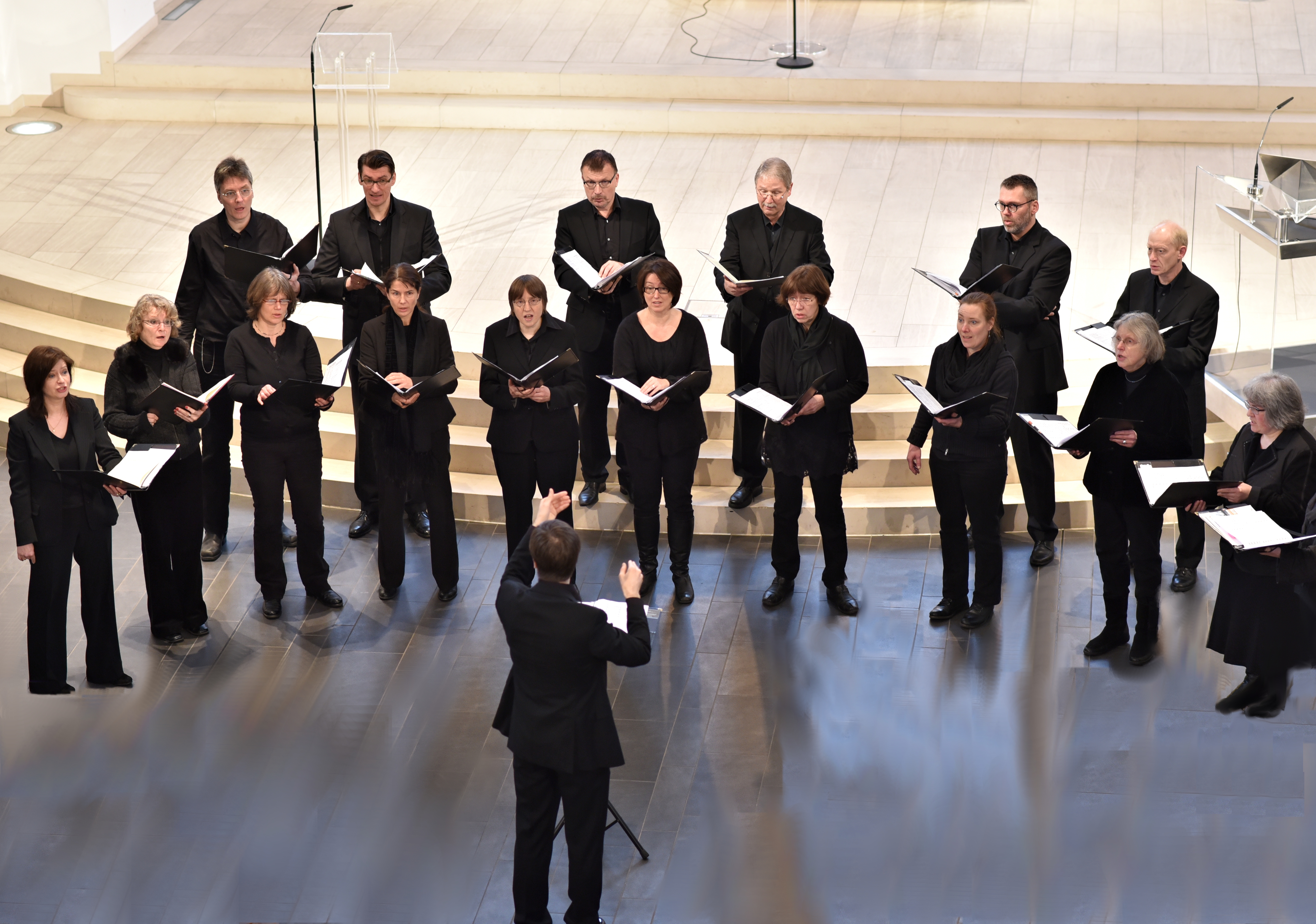 Vor dem Chorleiter, der nur in Rückenansicht zu sehen ist, stehen die Sängerinnen des Kammerchores in schwarzer Kleidung und singen aus schwarzen Mappen.